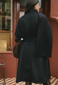 Woman wearing a dark coat with a belt, holding a brown bag, standing on a checkered floor outside a building with red walls and windows.