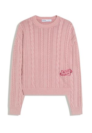 Pull en maille torsadée rose avec manches longues, poignets et ourlet côtelés, et un petit patch brodé sur le côté inférieur gauche.