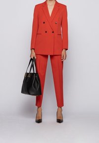 Veste rouge croisée à double boutonnage avec des boutons noirs et un pantalon assorti cintré. Associé à un sac fourre-tout noir texturé et des escarpins noirs pointus.
