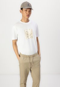Camiseta blanca con un cuadrado beige estampado en la parte delantera, combinada con pantalones jogger beige y una gorra gris claro. Corte relajado, tejido suave.