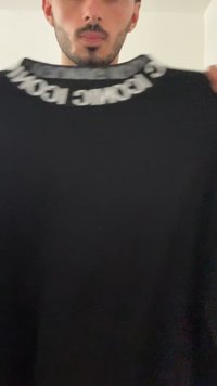 Felpa nera con collo a giro e tessuto testurizzato. La scritta bianca lungo il colletto recita "Prossimamente". Design semplice.