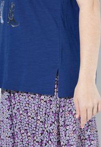 Modrá bavlněná blůza s krátkými rukávy a bočními rozparky, spárovaná s fialovou květinovou sukní s malými vzory a sklady.