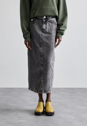 Person iført en grøn sweatshirt, grå acid-wash midi denim nederdel og gule slip-on sko, stående på en grå gulv mod en ensfarvet væg.