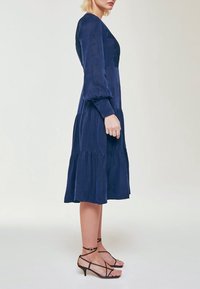 Kvinna som står i en marinblå långärmad midiklänning med lager på lager-skirt och svarta sandaletter med klack på en ljus bakgrund.