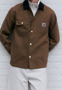 Giacca di tela marrone con colletto scuro, due tasche sul petto e due tasche laterali. Presenta bottoni a pressione dorati e una toppa con il logo del marchio.