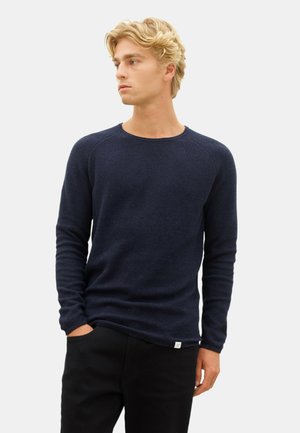 Navy knitted sweater with long sleeves, round neckline, and subtle texture. Paired with black pants, featuring a label at the hem.