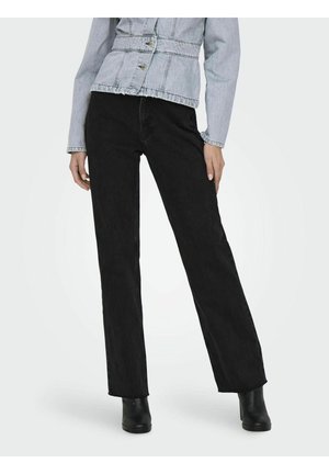 Frau trägt schwarze Jeans mit geradem Bein, eine helle Jeansjacke und schwarze Stiefeletten, steht vor einem einfarbigen hellen Hintergrund.