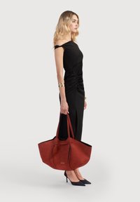 Punainen mokkasiinitotebag, jossa on mustia yksityiskohtia, tiivistetty yläosa, tukevat hihnat ja tilava sisus, jota mallilla on mustan mekon kanssa.