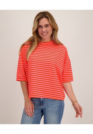 Vrouw met lang blond haar, die een koraalroze gestreept T-shirt met korte mouwen en een blauwe spijkerbroek draagt, glimlachend tegen een effen achtergrond.