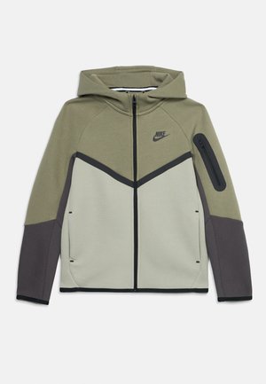Giacca Nike con cappuccio e cerniera frontale, parte superiore verde oliva, corpo inferiore grigio chiaro, maniche grigio scuro e tasca con zip sul braccio sinistro.