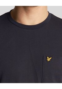 Muž nosící tmavě modré triko s kulatým výstřihem, na kterém je malé žluté vyšívané logo ptáčka na levé straně hrudi u kapsy.