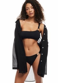 Sort bikini sæt med en bandeau-top med justerbare stropper og en matchende bund med guld hardware detaljer, parret med en gennemsigtig sort skjorte.
