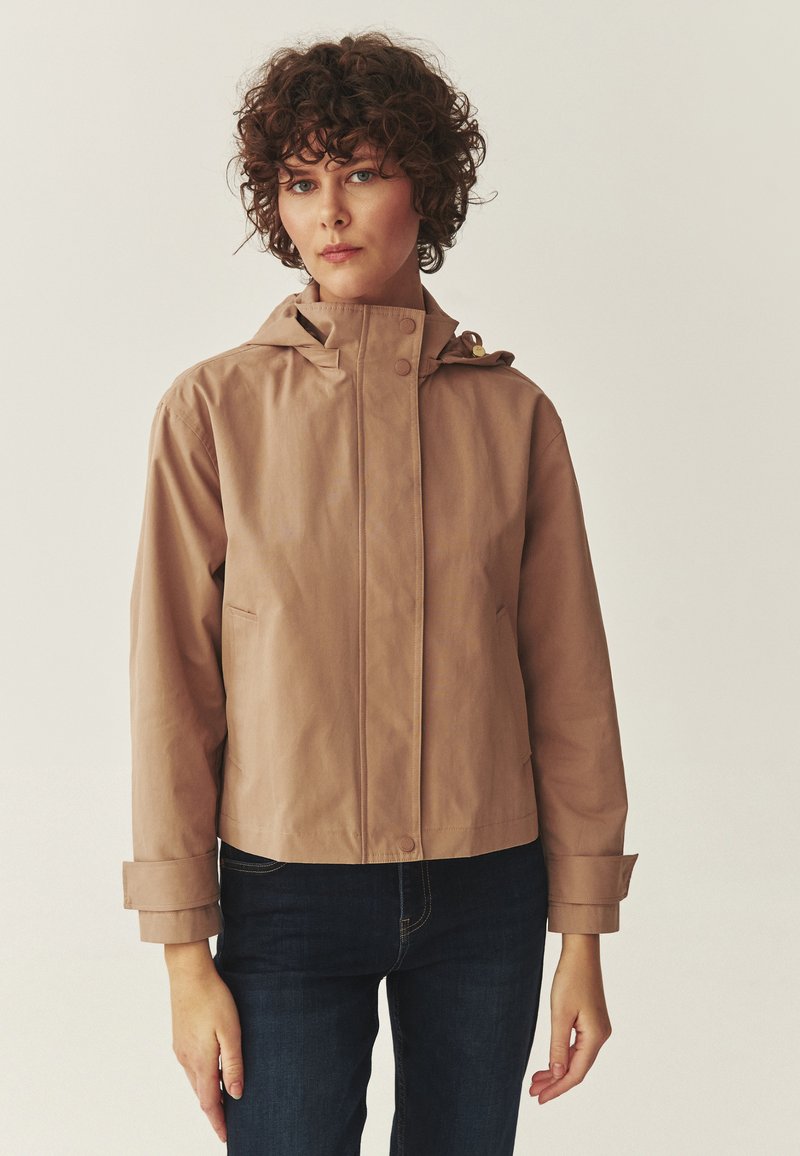 Femme aux cheveux bouclés bruns portant une veste à capuche beige et un jean bleu foncé sur un fond clair uni.