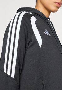 Μαύρο φούτερ με κουκούλα από μαλακό ύφασμα, με τρεις λευκές ρίγες στον ώμο και μικρό λογότυπο. Διαθέτει τσέπη με φερμουάρ.