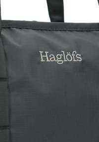 Borsa tote in tessuto nero con superficie testurizzata, cuciture in evidenza e il nome del marchio "Haglöfs" ricamato in bianco.