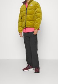 Grön puffjacka med dragkedja, ribbade muddar; lager-på-lager över en rosa skjorta; kombinerad med svarta byxor och vinröda träningsskor.