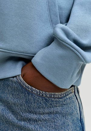 Vaaleansininen svetari, jossa on resorit hihansuissa, yhdistetty tummansinisiin farkkuihin. Kankaassa on sileä pinta ja näkyvät ompelusaumat.