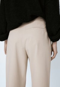 Persona che indossa pantaloni beige e una maglietta nera a maniche lunghe con texture, vista da dietro all'altezza della vita.