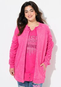 Chaqueta rosa con cremallera y mangas fruncidas, presenta brillantes acentos plateados. Debajo, una camiseta rosa a juego con texto de rhinestones. Combinada con jeans desgastados.