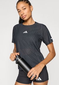 Czarna krótka sportowa koszulka z teksturowanym wzorem i białym logo Adidas; w zestawie z czarnymi szortami i trzyma czarną, elegancką butelkę na wodę.