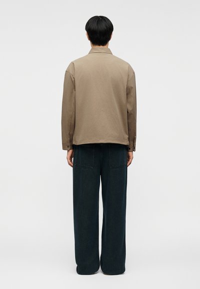 Άτομο με κοντά μαύρα μαλλιά στέκεται με την πλάτη, φορώντας μπεζ σακάκι και χαλαρό μαύρο παντελόνι, με φόντο ένα απλό λευκό τοίχο.