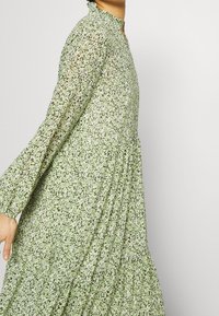 Vaaleanvihreä kukallinen mekko, jossa on korkea kaulus, pitkät hihat ja liehuva, laskostettu muotoilu. Painettuna kuvioina pieniä, hajallaan olevia kukkia.