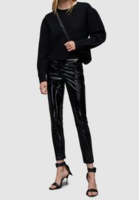 Schwarzer Pullover mit Puffärmeln, kombiniert mit glänzenden schwarzen Kunstlederhosen und schwarzen Riemchen-High-Heels. Eine schlichte Umhängetasche.