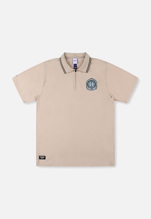 Polo beige à manches courtes avec un col zippé, des accents rayés noirs et blancs, et un logo circulaire brodé sur le côté gauche de la poitrine.