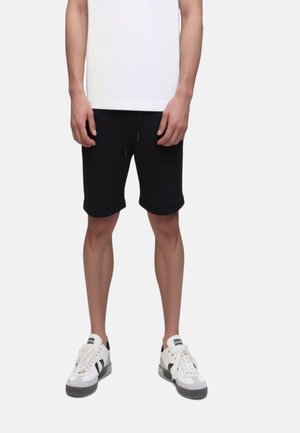 Hombre con pantalones cortos negros con cordón, camiseta blanca y zapatillas blancas con detalles en negro, de pie contra un fondo liso.