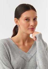 Grijze gebreide trui met een V-hals en geribbelde manchetten. Model draagt zilveren oorbellen in de vorm van een ring, met een gladde afwerking en ronde vorm.