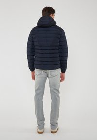 Giacca puffer navy con cuciture orizzontali, cappuccio e polsini elastici, abbinata a jeans chiari e scarpe casual, vista da dietro.