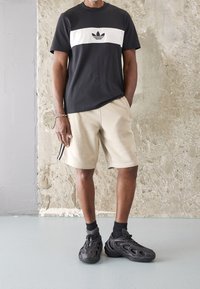 Černobílé tričko Adidas s logem na hrudi, spárované s béžovými kraťasy s černým pruhem. Nosené s černými texturovanými teniskami.