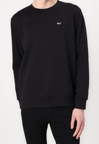 Persona con un sudadera negra de manga larga lisa que tiene un pequeño logo rojo, blanco y azul en el lado izquierdo del pecho, combinado con pantalones negros.