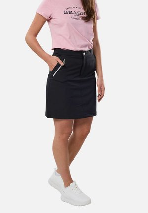 Kobieta w różowym t-shircie, czarnej spódnicy na zamek błyskawiczny i białych trampkach, stoi z jedną ręką w kieszeni spódnicy na białym tle.