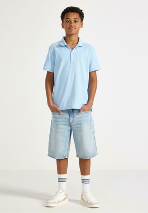 Jongen draagt een lichtblauw poloshirt, knielange denim shorts, witte hoge sokken met strepen en witte sneakers, staat met handen in de zakken.