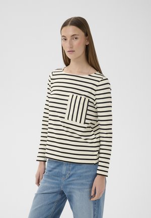 Kobieta ubrana w czarno-białą koszulkę z długim rękawem w poziome paski z pionowo paskowaną kieszenią i jasnoniebieskie dżinsy, stojąca na tle białej ściany.