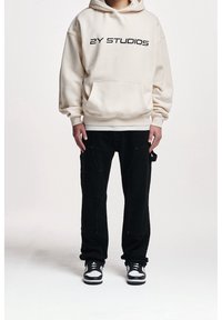 Kermansweater dengan teks hitam "2Y STUDIOS", dipadukan dengan celana cargo hitam yang memiliki saku. Dipakai dengan sepatu sneakers hitam dan putih.