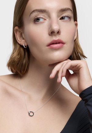 Giovane donna con capelli castano chiaro corti che indossa un orecchino a cerchio d'argento e una delicata collana d'argento con un ciondolo circolare nero.