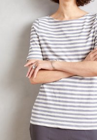 Camiseta de manga corta a rayas en gris claro y blanco de algodón, con escote barco y corte relajado. Brazos cruzados al frente, mostrando un anillo de plata y pulseras.