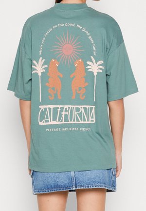 Groen oversized T-shirt met een retro grafisch ontwerp van twee oranje luipaarden, palmbomen, een zon en de tekst "CALIFORNIA" eronder.