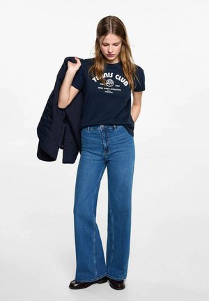 T-shirt in cotone navy con grafica "Tennis Club", abbinato a jeans a vita alta svasati di colore azzurro chiaro, indossato con una giacca scura su un braccio.