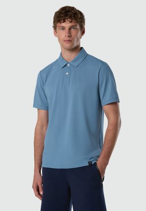 Giovane uomo con capelli ricci che indossa una polo a maniche corte azzurro chiaro e pantaloni scuri, in piedi con una mano in tasca.