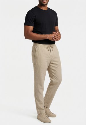 Uomo con maglietta nera a maniche corte, pantaloni beige con coulisse e sneakers beige in piedi con le mani incrociate.