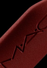 Matowy burgundowy produkt do ust z wytłoczonym logo i wzorem w zygzak. Gładka tekstura z delikatnym połyskiem, skośny koniec do precyzyjnej aplikacji.