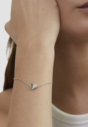 Silberarmband mit herzförmigem Anhänger, glatter Oberfläche und Gliederkette, getragen an einem Handgelenk vor neutralem Hintergrund.