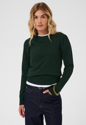 Femme blonde portant un pull vert foncé à manches longues sur un haut blanc, un jean bleu, posant avec une main dans la poche devant un fond gris.