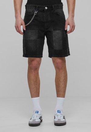 Pantalones cortos de mezclilla negra con un diseño de patchwork, detalles desgastados y un acento de cadena plateada. Combinados con calcetines deportivos blancos y zapatillas.