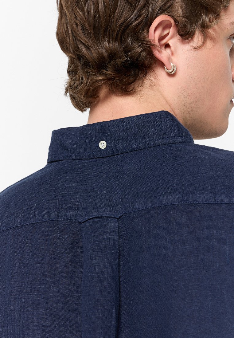 Hombre con cabello castaño que lleva un pendiente de aro plateado y una camisa de button-down azul marino, mostrado desde atrás y de perfil lateral.