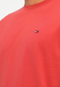 Červené bavlněné tričko s kulatým výstřihem a malým vyšitým logem v námořnické modři, bílé a červené barvy na levé straně hrudi.