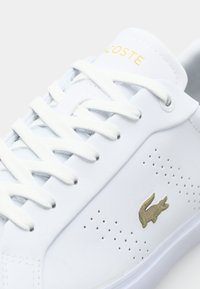 Baskets en cuir blanc avec design perforé. Elles sont dotées de lacets blancs, d'un logo doré sur la languette et d'un emblème de crocodile doré sur le côté.